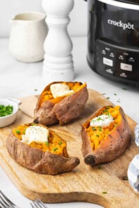 3 open baked sweet potatoes with a pat of butter on top and a crockpot in the background.