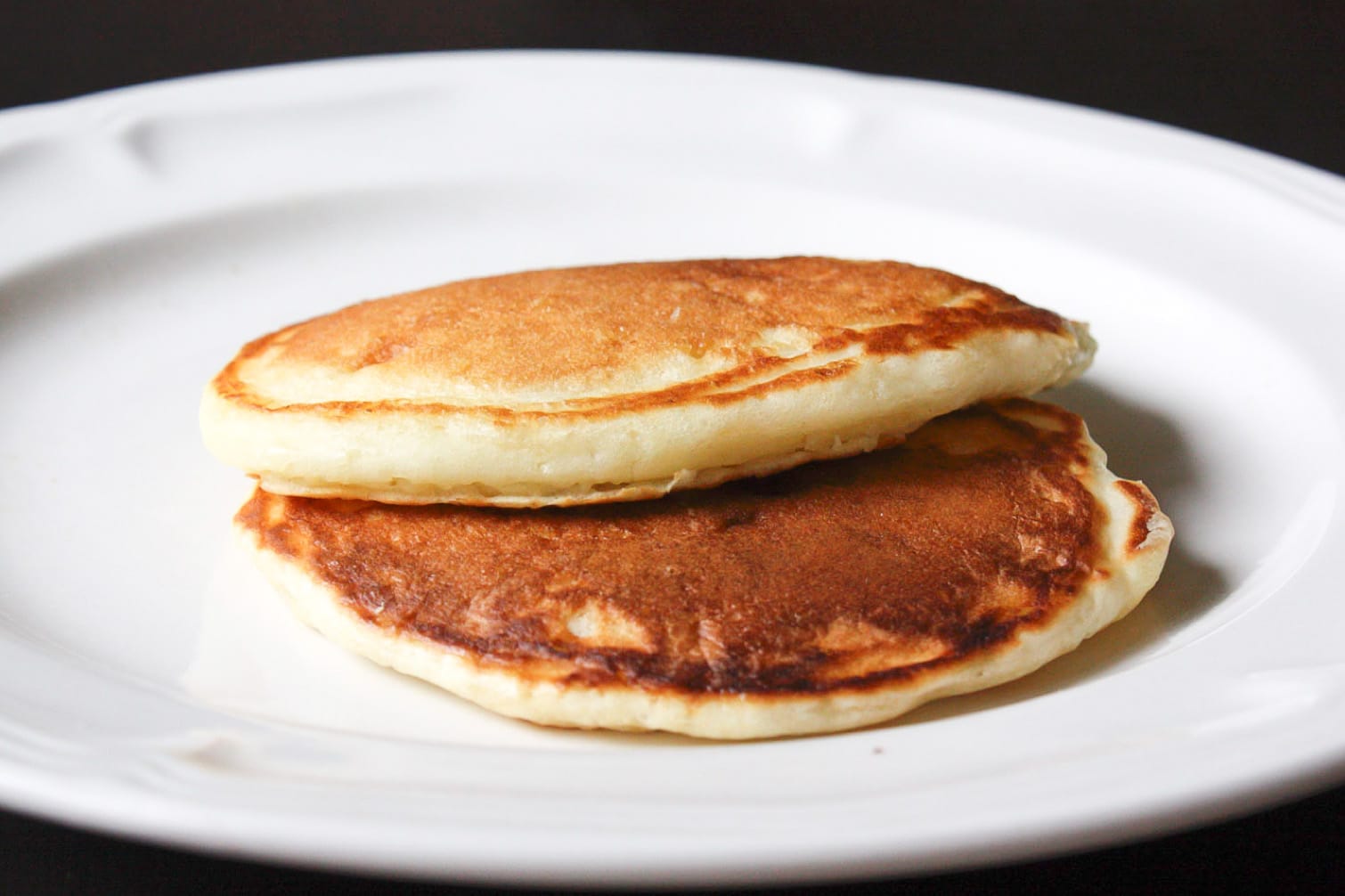 2 golden buttermilk pancakes stacked together on a white plate with a black background