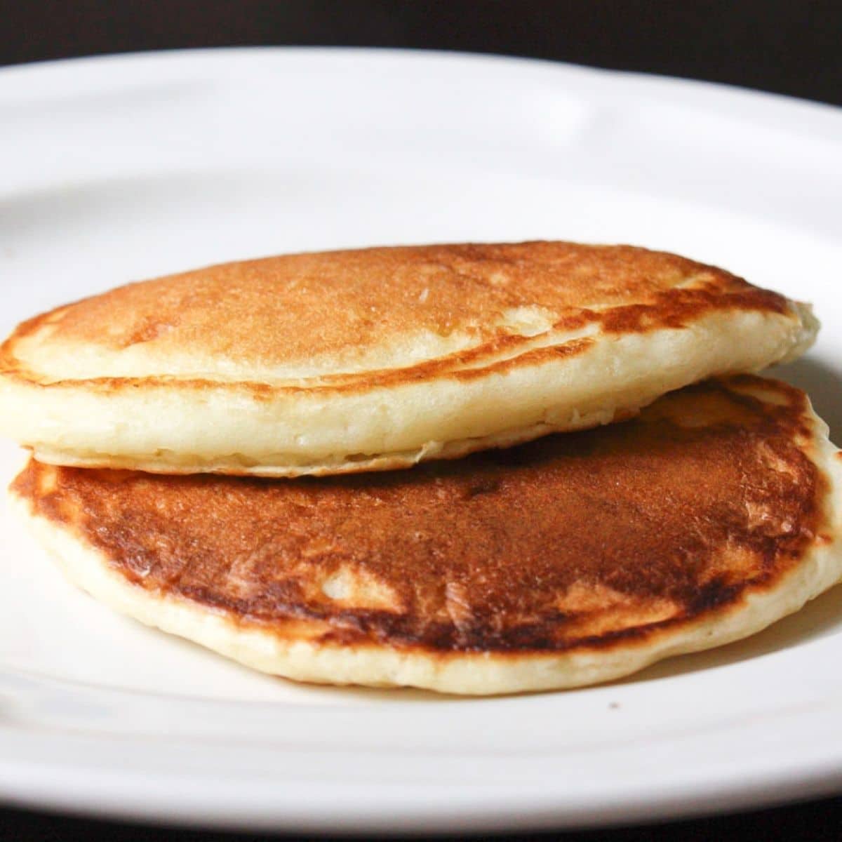 2 golden pancakes stacked together on a white plate with a black background