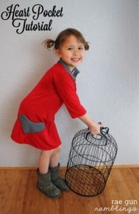 little girl in red long sleeve dress with gray heart holds black wire bird cage. White all and wood floor in background