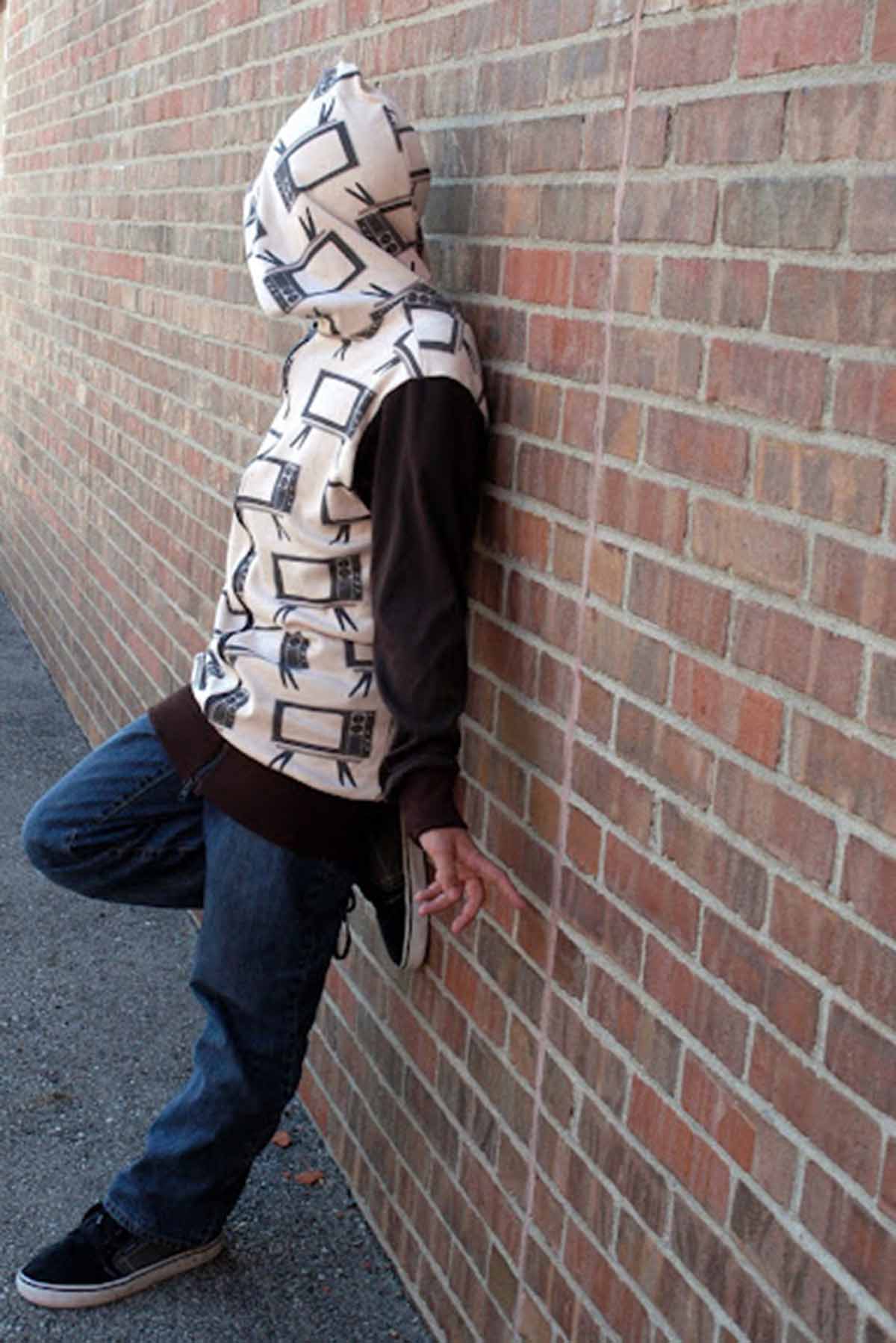 Boy leaning against a brick wall with a hoodie over his head.