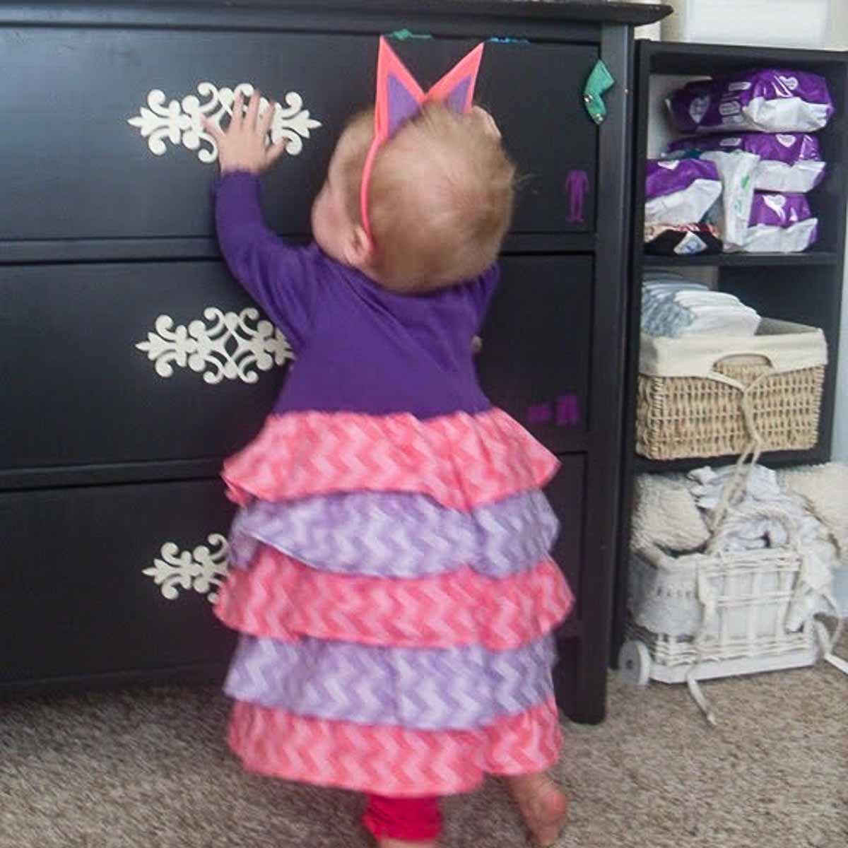Baby in a ruffled shirt dress holding onto a dresser.