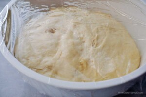 Risen yeast dinner roll dough in a bowl covered in plastic wrap.