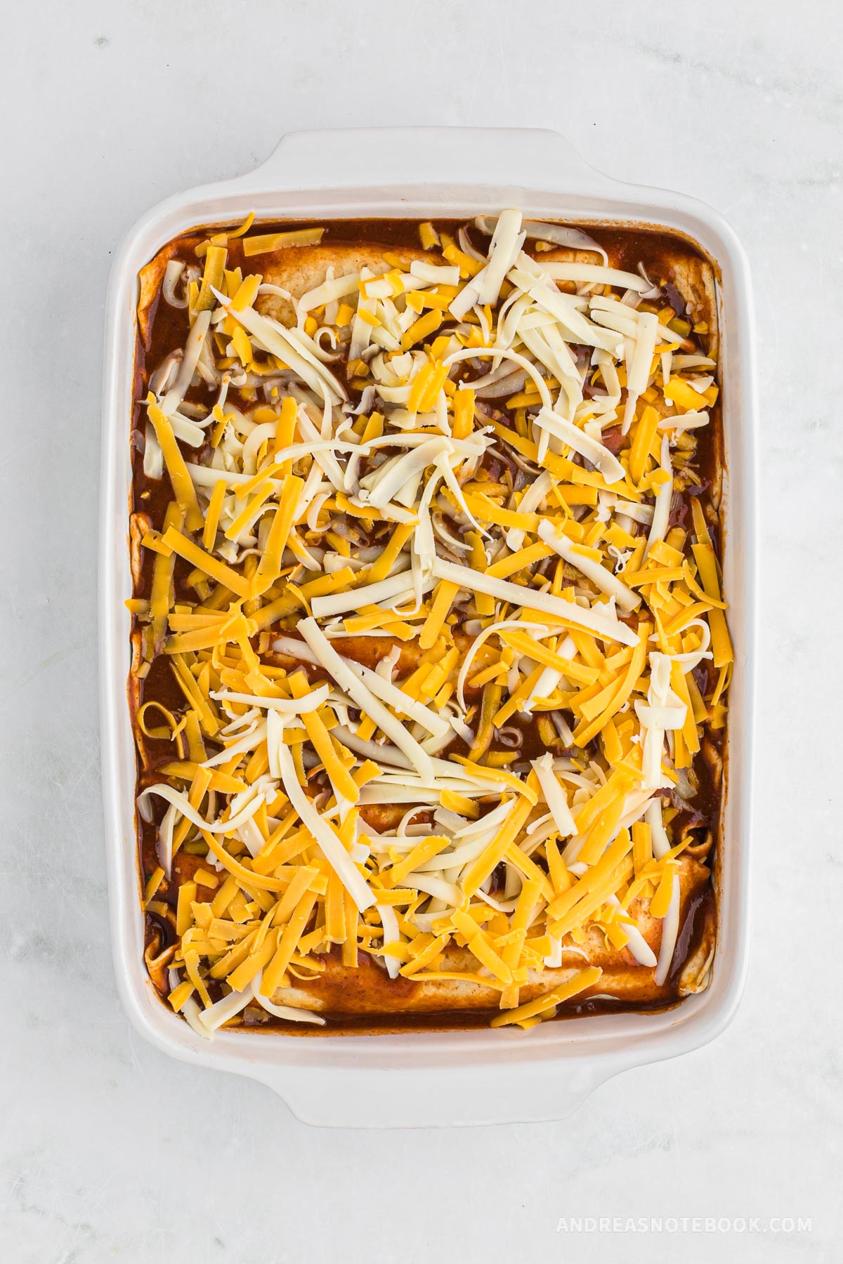 Enchiladas rolled up and placed into a pan. Uncooked shredded cheese on top.