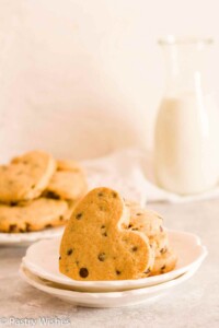 Two white plates with heart shaped chocolate chip cookies in front of a glass of milk.