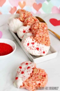 Pink heart shaped Rice Krispie treats with the left half dipped in white chocolate and sprinkled with valentines sprinkles.