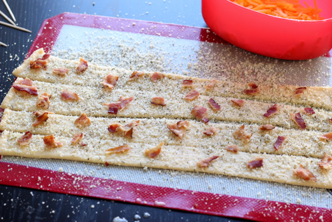 Uncooked strips of dough with seasoning and bacon on top.