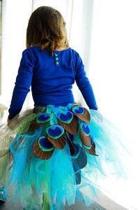 Girl wearing blue shirt and blue tutu with sewn felt peacock feathers.