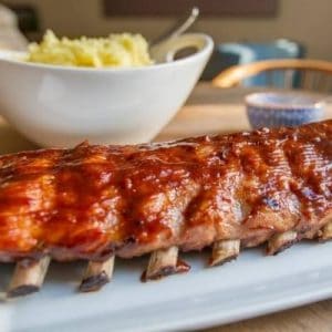 barbecue sauce on rack of ribs with bowl of mashed potatoes in background