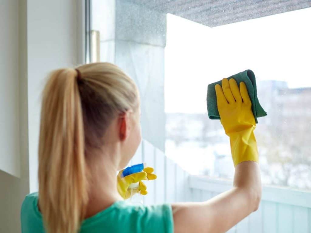 woman with blonde pony tail uses yellow rubber gloves and green cloth to wipe large window