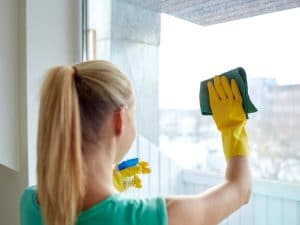 woman with blonde pony tail uses yellow rubber gloves and green cloth to wipe large window