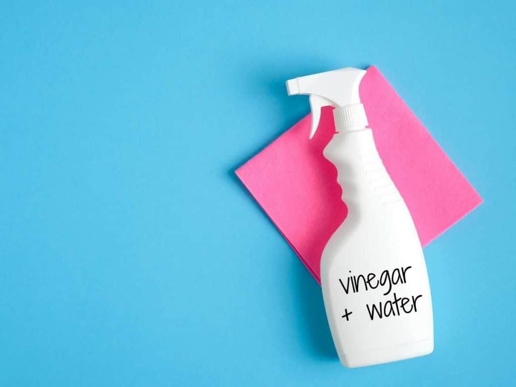 blue background. pink microfiber towel and white spray bottle with vinegar cleaning solution. Bottle says "vinegar + water"