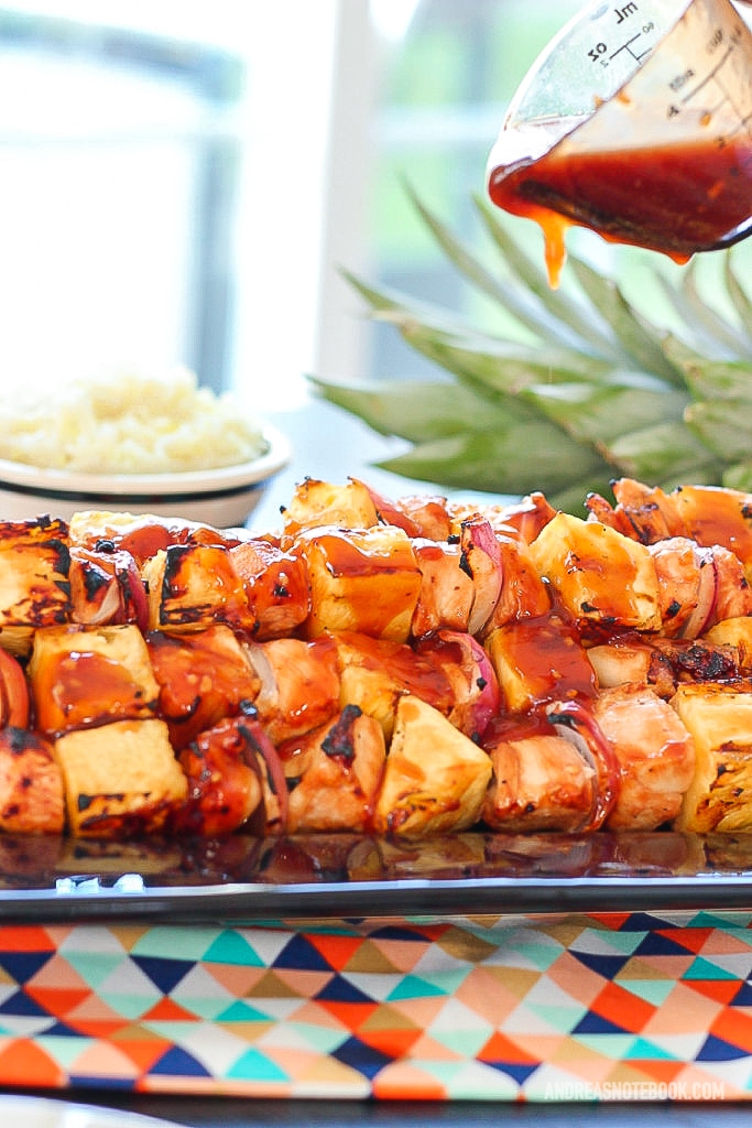 black rectangular plate with a stack of kabab skewers with pineapple, pineapple and onion with a glaze on top being dripped onto kabobs from a measuring cup from above