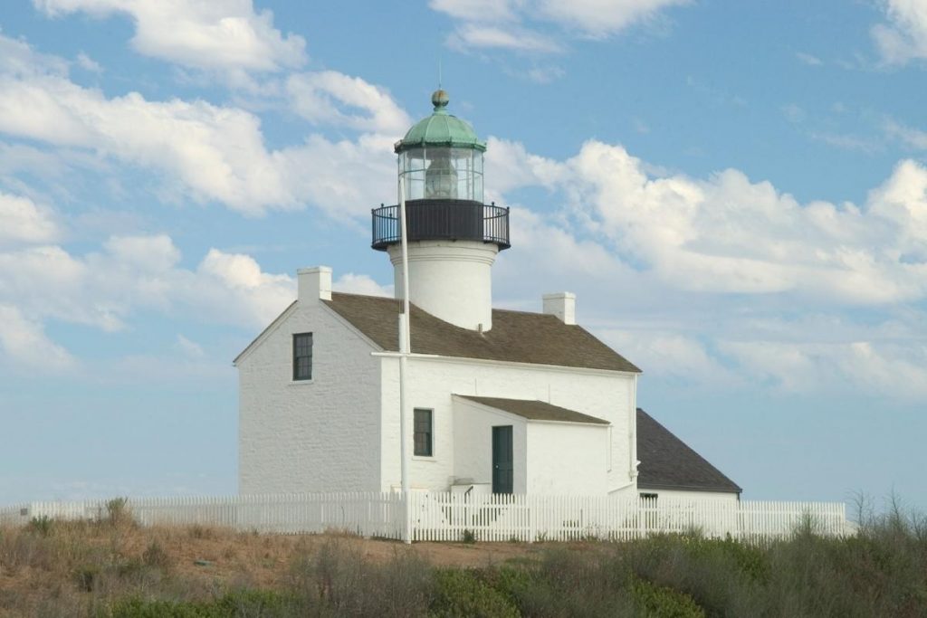 Old point loma lighthouse things to do in san diego - White lighthouse sits on dirt with seagrass around. The one story building with lighthouse above has a short white fence surrounding. Sky is blue with white clouds.