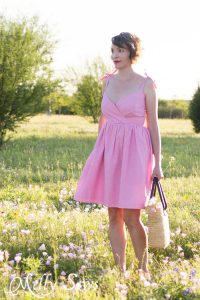 White woman standing in green field wearing a pink sundress with wrap front detail and thin straps holding a handbag
