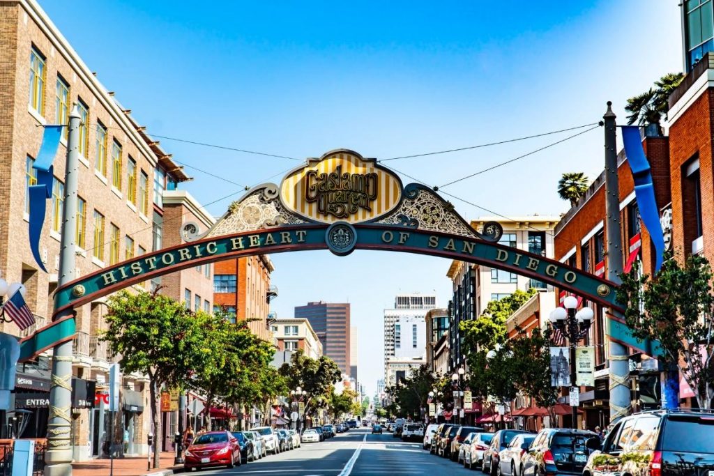 The Gaslamp Quarter sign arching over the street is the beginning of the gaslamp quarter. The tourist attraction is one of the best things to do in San Diego. Four to five story buildings on each side of the street with cars parked all along the street.