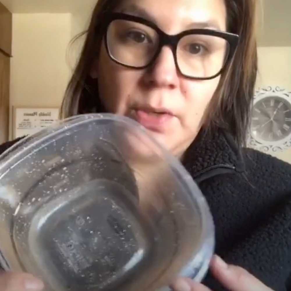 White woman with black glasses, brown straight hair and a black fleece teaches how to clean orange stains off plastic containers. She holds up stained container to show it off.