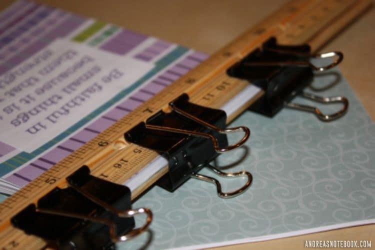 Two wood rulers and three black binder clips clamping down a stack of papers with decorative paper on top in photo of how to make a notepad. Clamps used to hold DIY notepad together while the glue for binding notepads (padding compound) dries.