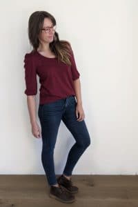woman stands on wood floor against a white wall. She's wearing skinny blue jeans and a burgundy t-shirt with banded half sleeve puffy sleeves