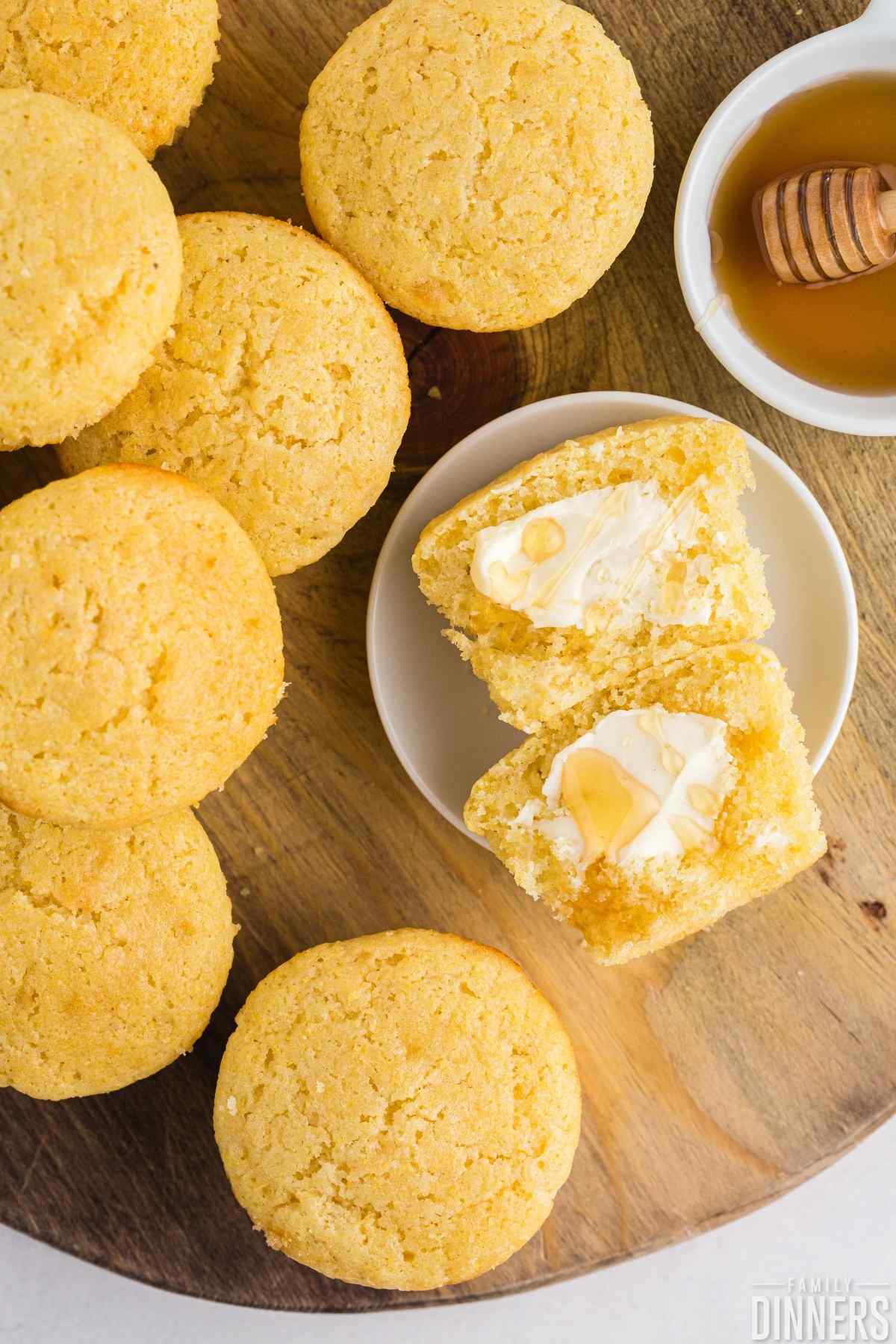 Cutting board with stack of cornbread muffins including one split open with butter and honey on top.