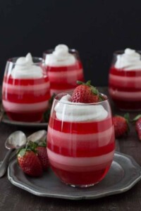 4 stemless wine glasses full of pink and red layered jello cups topped with whipped cream and fresh strawberry.