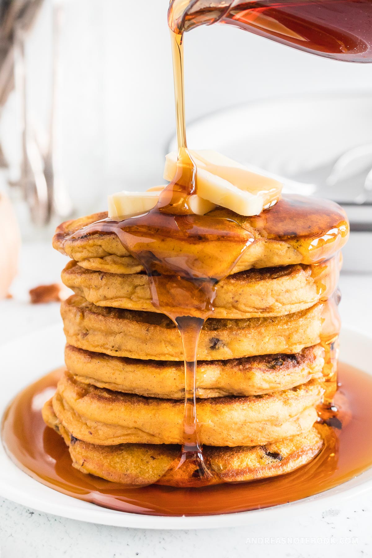 Syrup pouring on large stack of pumpkin pancakes with chocolate chips and pecans.