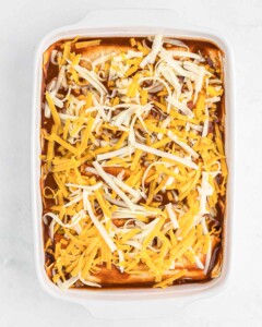 Casserole dish full of enchiladas covered in red enchilada ranchero sauce and shredded cheese.