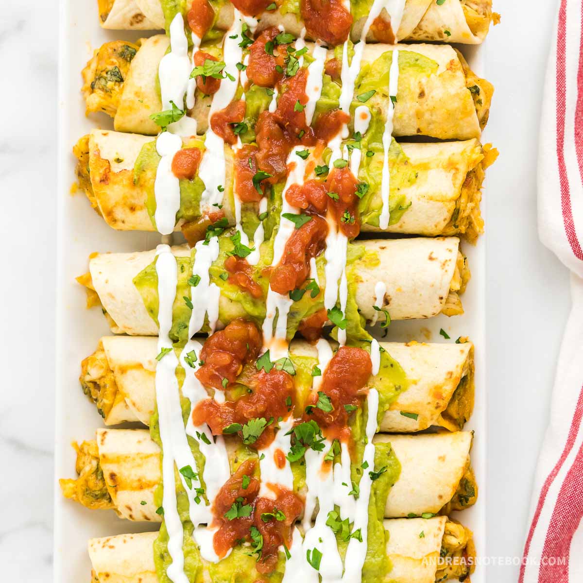 Platter with 7 cooked chicken taquitos in a row covered in salsa, guacamole and sour cream.