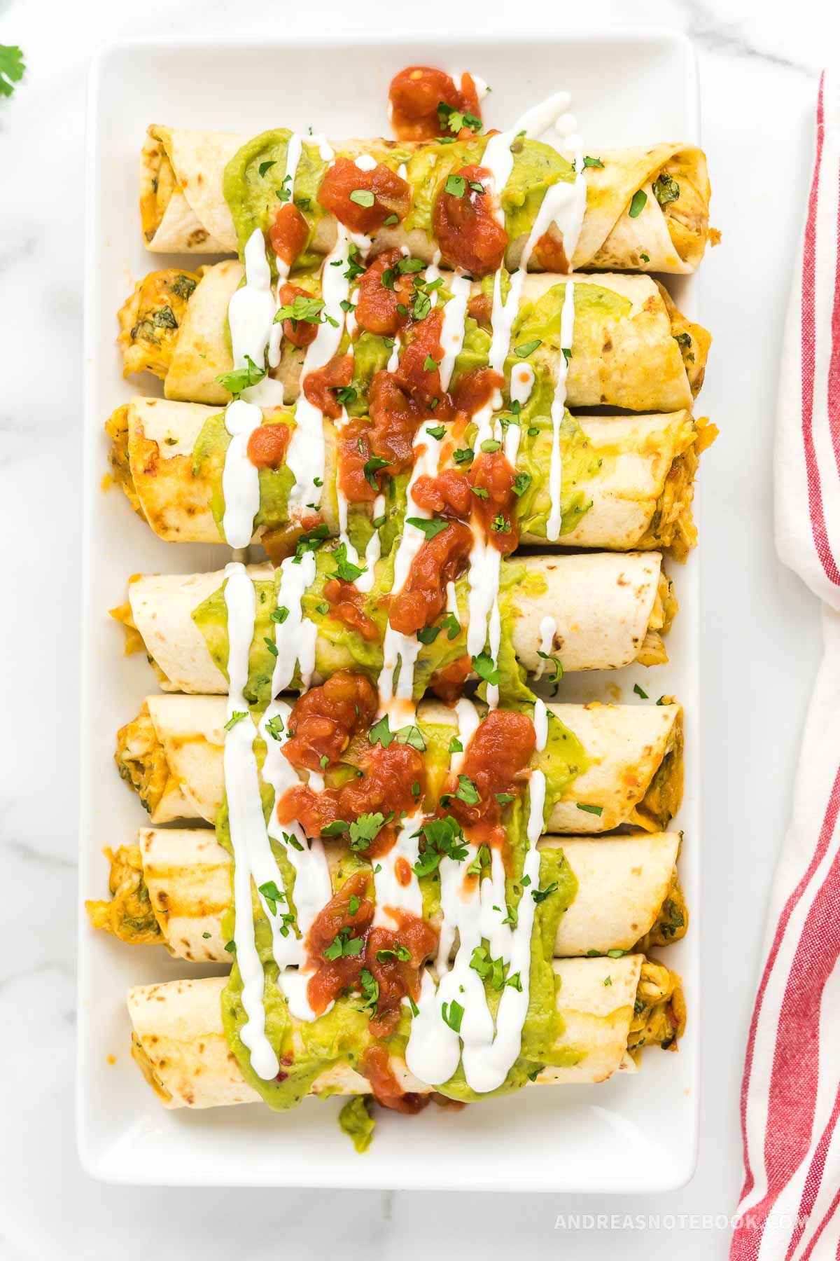 Platter with 7 cooked chicken taquitos in a row covered in salsa, guacamole and sour cream.