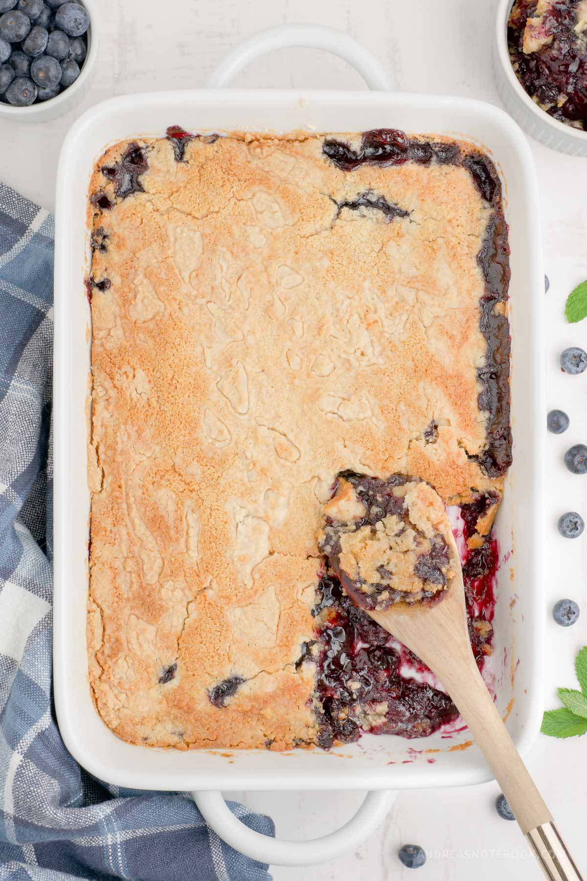 Cooked blueberry dump cake in a baking dish with a wooden spoon scooping out.
