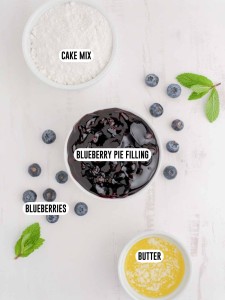 Blueberry dump cake ingredients in bowls including cake mix, blueberry pie filling, melted butter and fresh blueberries.