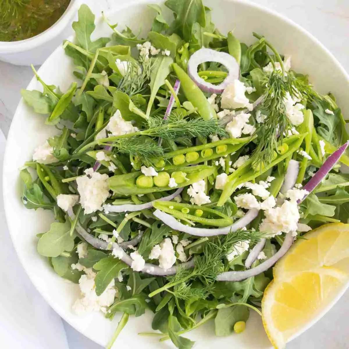 Snap pea and arugula salad in a white bowl with a lemon wedge.