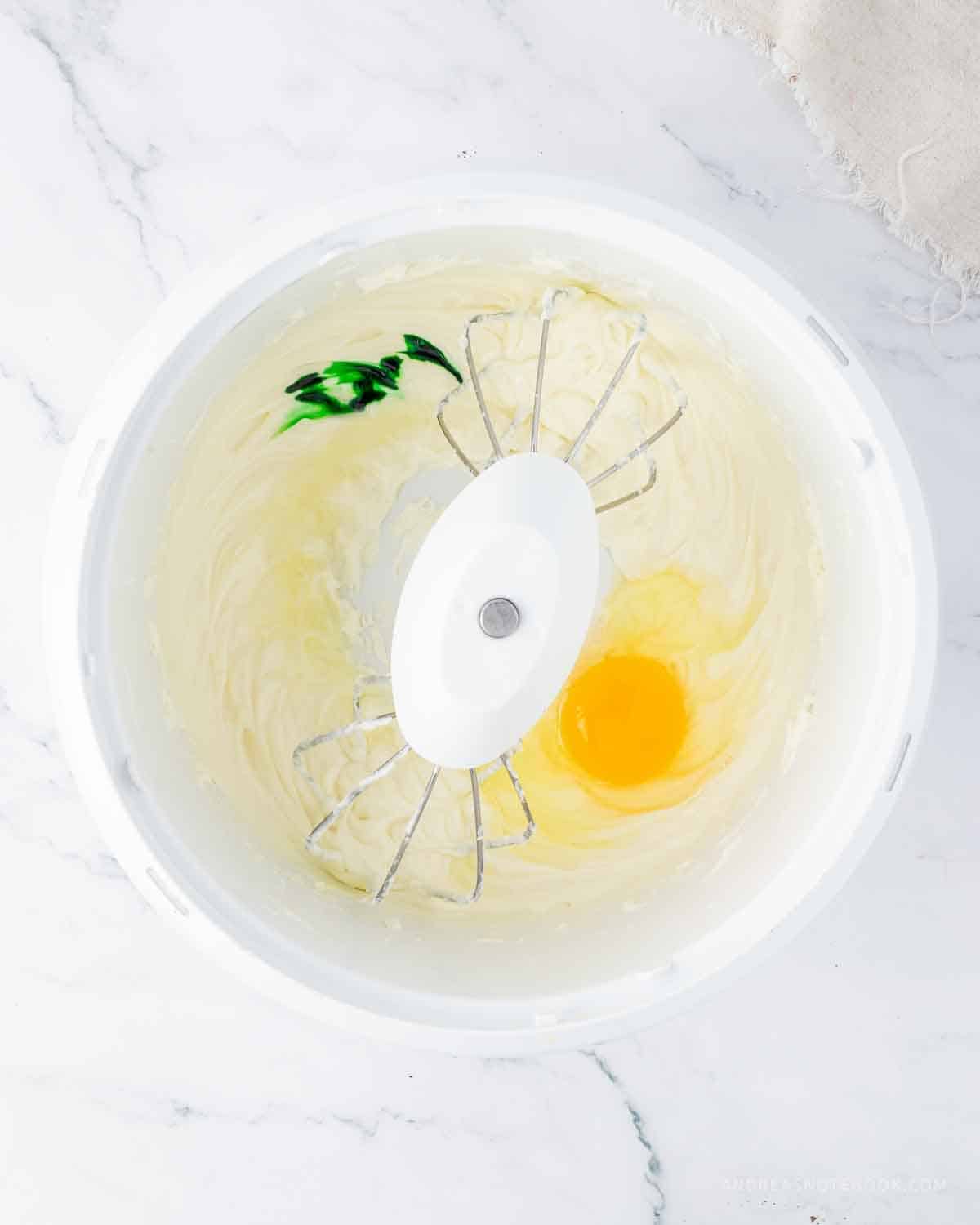 Green food coloring and egg added to cream cheese and sugar in the mixing bowl.