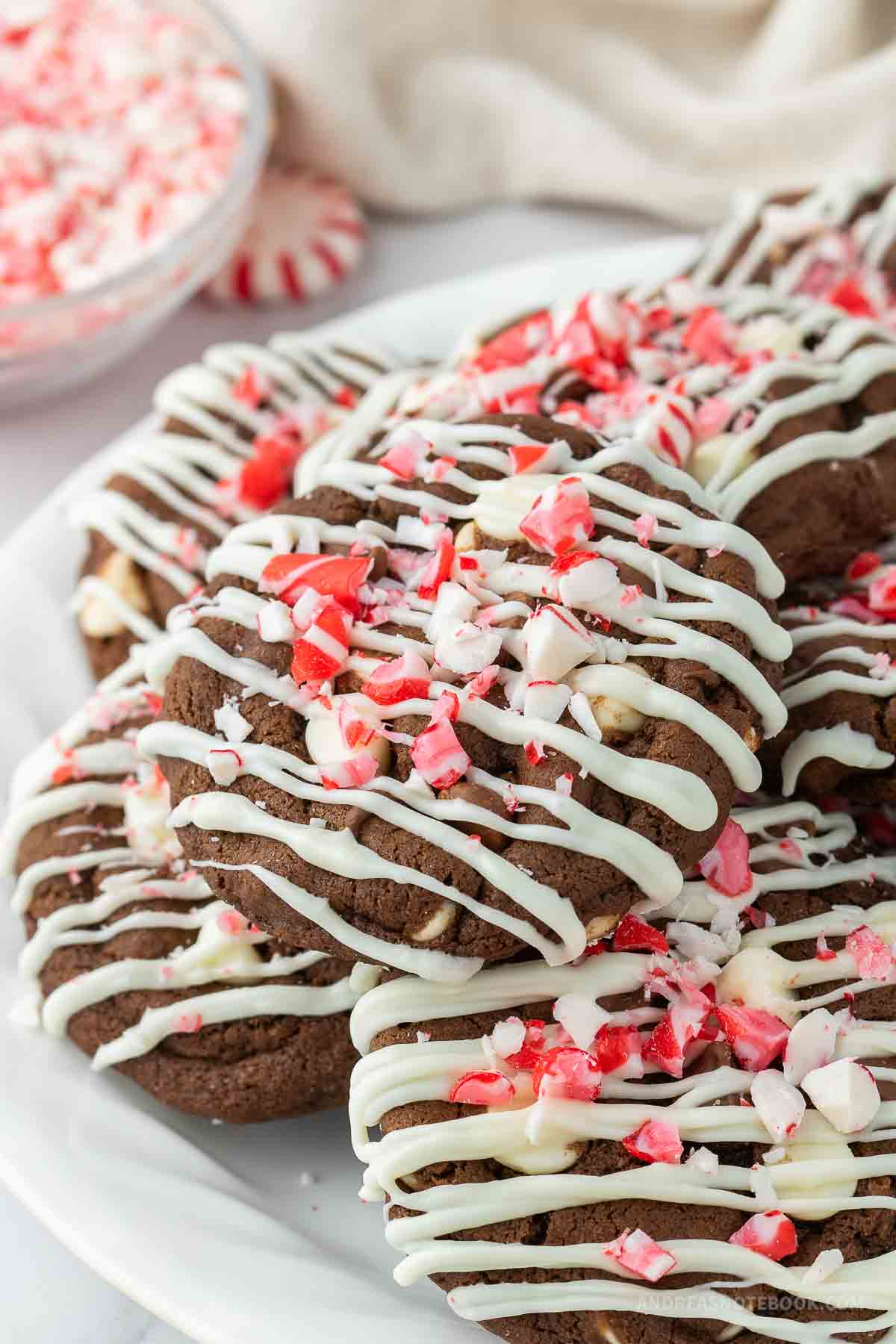 Pile of double chocolate peppermint cookies on a plate with white chocolate drizzle and crushed candy cane on top.