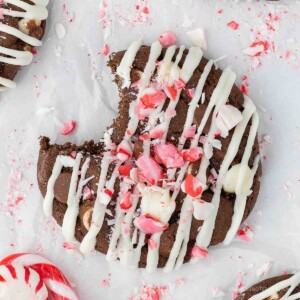 Double chocolate peppermint bark cookie with white chocolate drizzle and candy cane crumbles on top and a bite out of it.