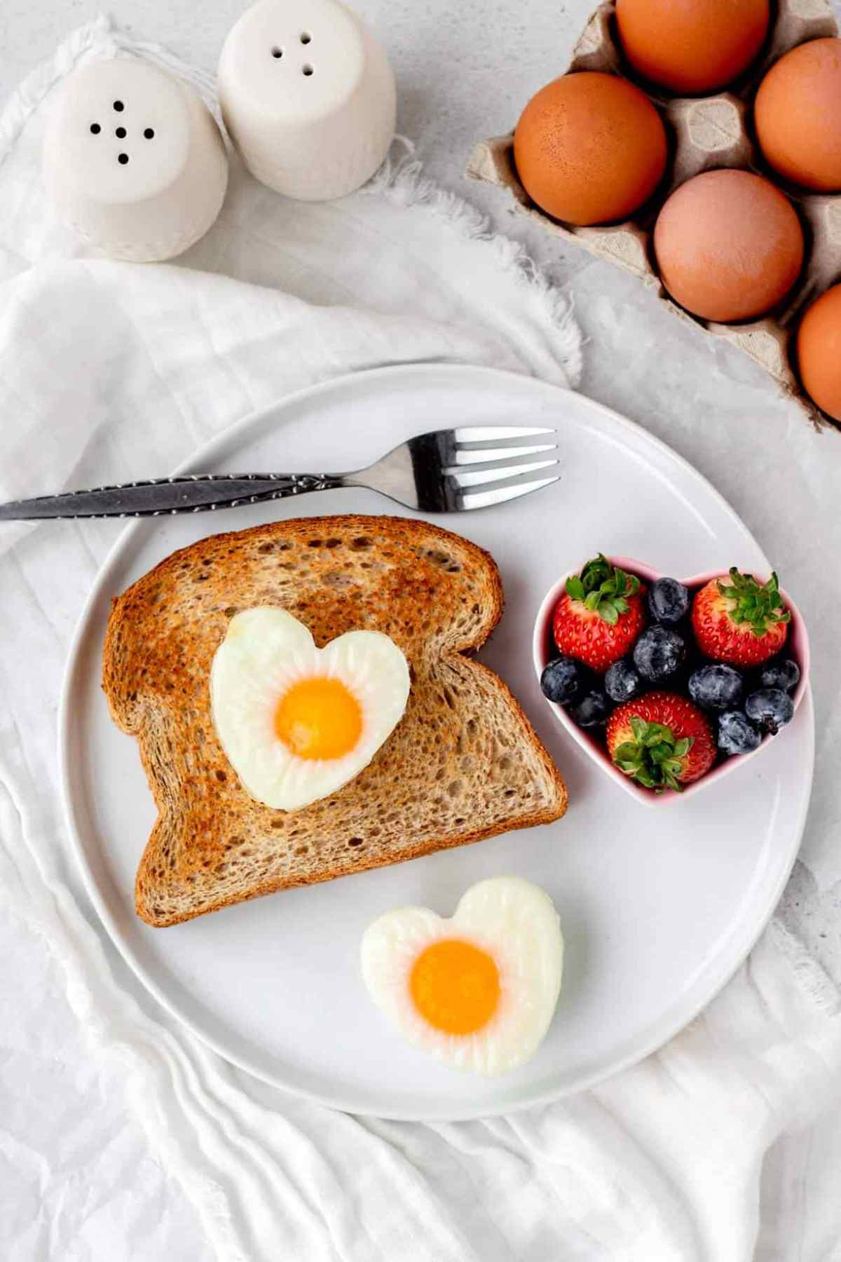 Heart shaped baked eggs on a piece of toast next to a heart shaped cookie cutter full of fresh berries on a plate.