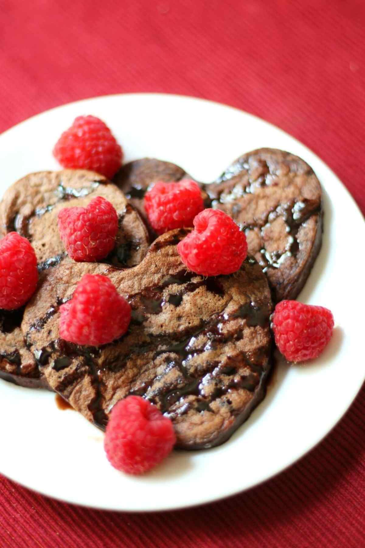 Plate full of heart shaped brown chocolate pancakes with fresh raspberries.