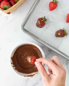 Strawberry being dipped into a bowl of chocolate and placed on a baking sheet.