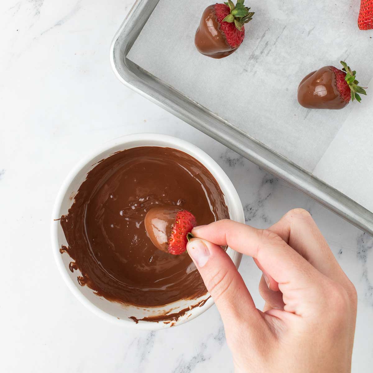 Hand dipping a strawberry in melted chocolate, two dipped strawberries on parchment paper on a baking sheet.