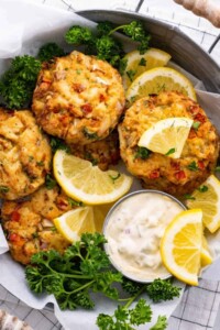 Serving dish full of lump crab cakes with lemon wedges and small container or white sauce.