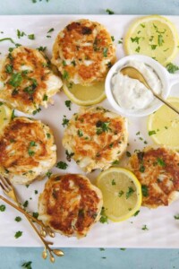 6 crab cakes on serving platter with sliced lemon, sauce and fresh parsley.