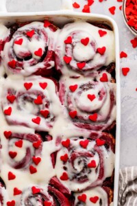 Baking pan full of red velvet cinnamon rolls topped with red hearts and icing.