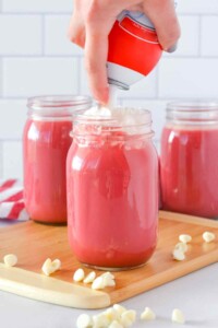 Mason jars full of thick pink red velvet hot chocolate with whipped cream being sprayed on top.