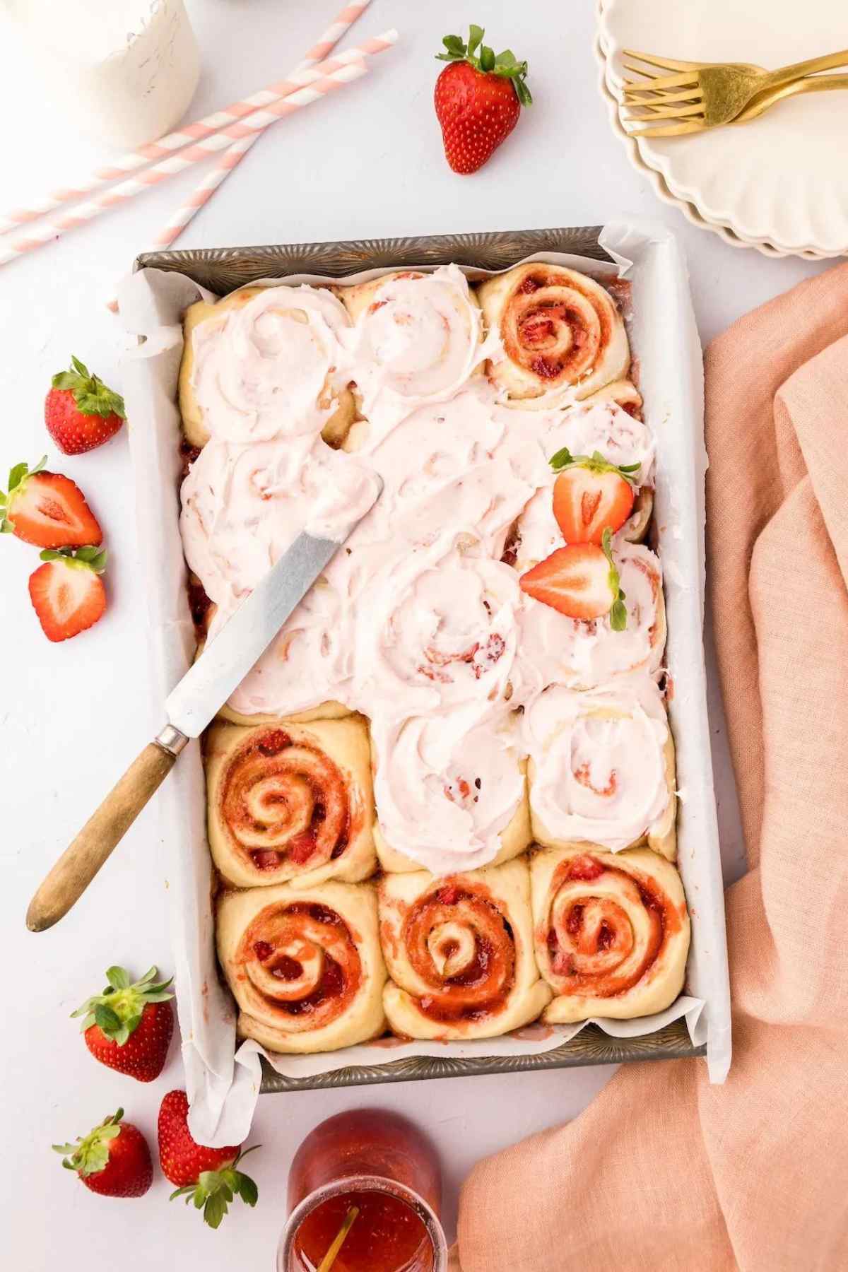 Casserole baking dish full of 12 strawberry cinnamon rolls being iced with pink icing.