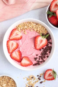 White bowl full of pink strawberry smoothie with sliced strawberries, mini chocolate chips and granola on top.