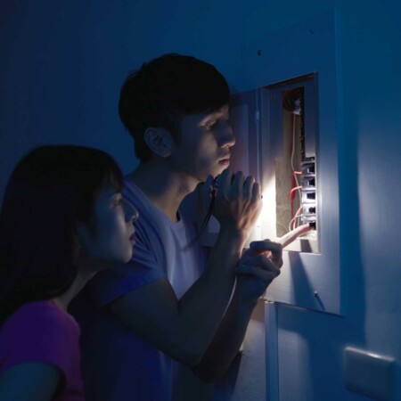 two people looking at an electrical panel with a flashlight in the dark.